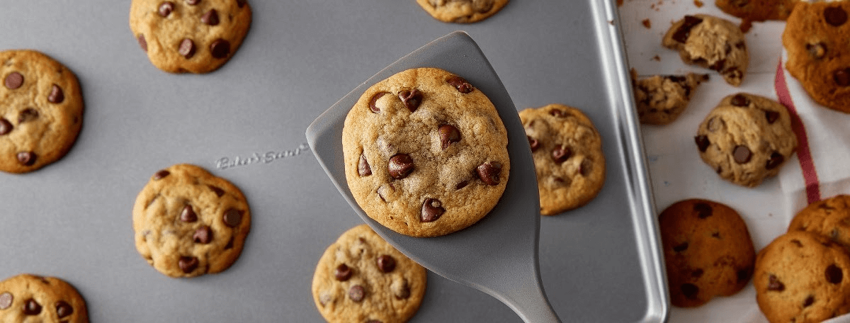 Golden chocolate chip cookies on a Baker's Secret cookie sheet with a spatula