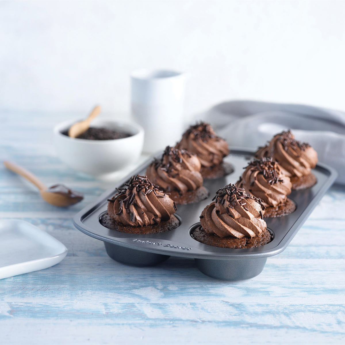 Chocolate frosted cupcakes in a 12-cup muffin pan on a light blue table